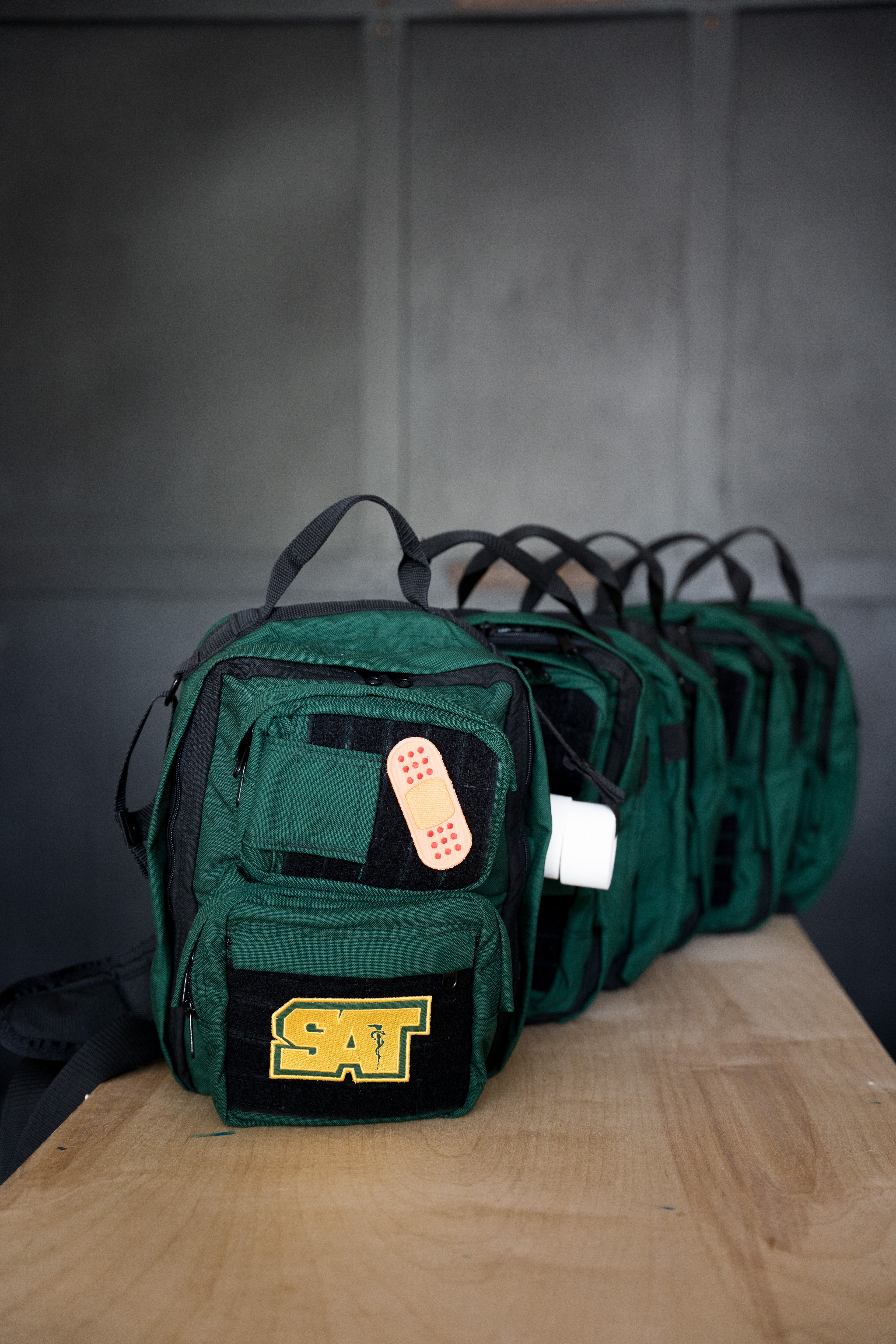 A row of dark green tactical-style backpacks with black straps and a yellow 'SAT' patch on the front, displayed on a wooden table against a dark gray paneled wall.