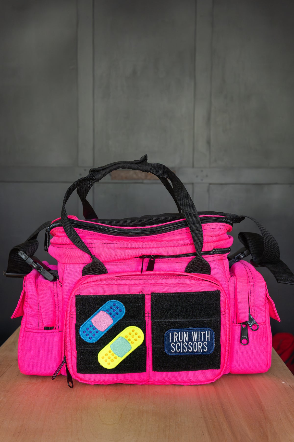 A bright pink utility bag with black handles and front pockets, featuring two band-aid shaped patches and a patch that reads 'I RUN WITH SCISSORS', displayed on a wooden surface against a gray paneled background.